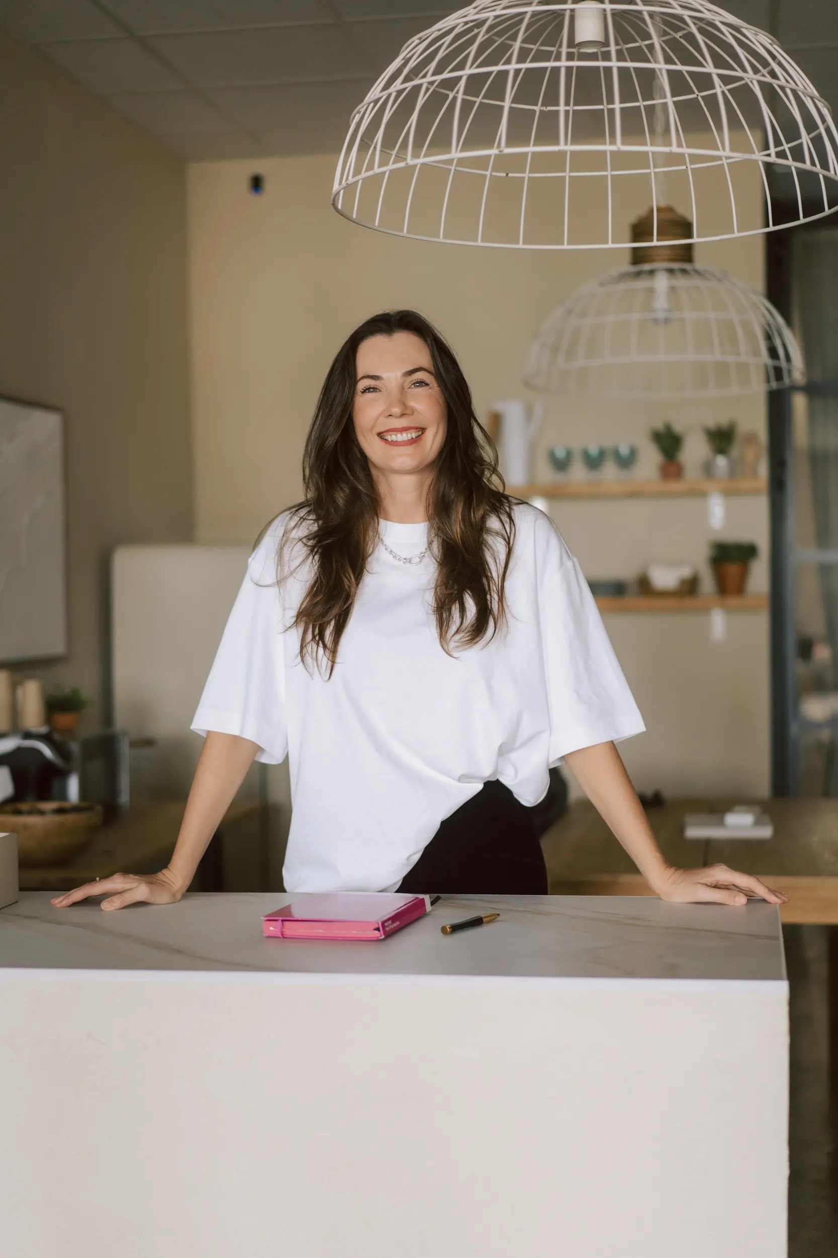 Mujer sonriente de cabello largo y suelto, vestida con una camiseta blanca, de pie en un escritorio moderno. En la mesa hay una libreta rosa y un bolígrafo, con un ambiente acogedor y decorado con plantas en el fondo.