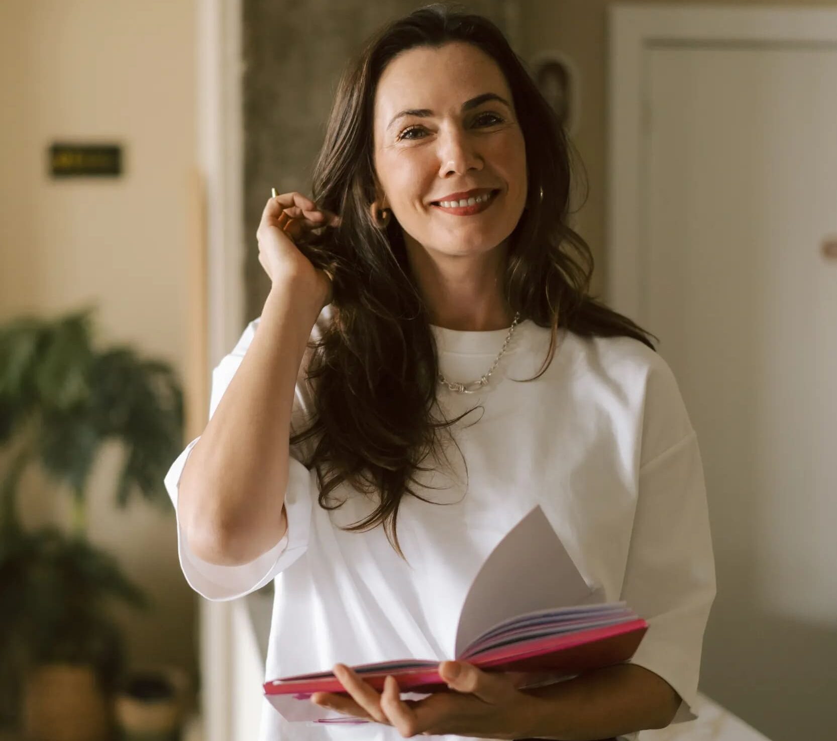 Mujer sonriente con cabello largo y oscuro, sosteniendo un libro abierto, vestida con una camiseta blanca, en un ambiente acogedor con decoración minimalista y plantas de interior.